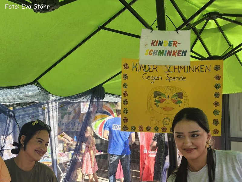 Zwei Frauen sitzen unter einem grünen Vordach an einem Schminkstand für Kinder. Über ihnen hängt ein buntes Schild mit einem gemalten Gesicht und der Aufschrift „Kinder Schminken gegen Spende“. Im Hintergrund sind Kinder und Eltern zu sehen.
